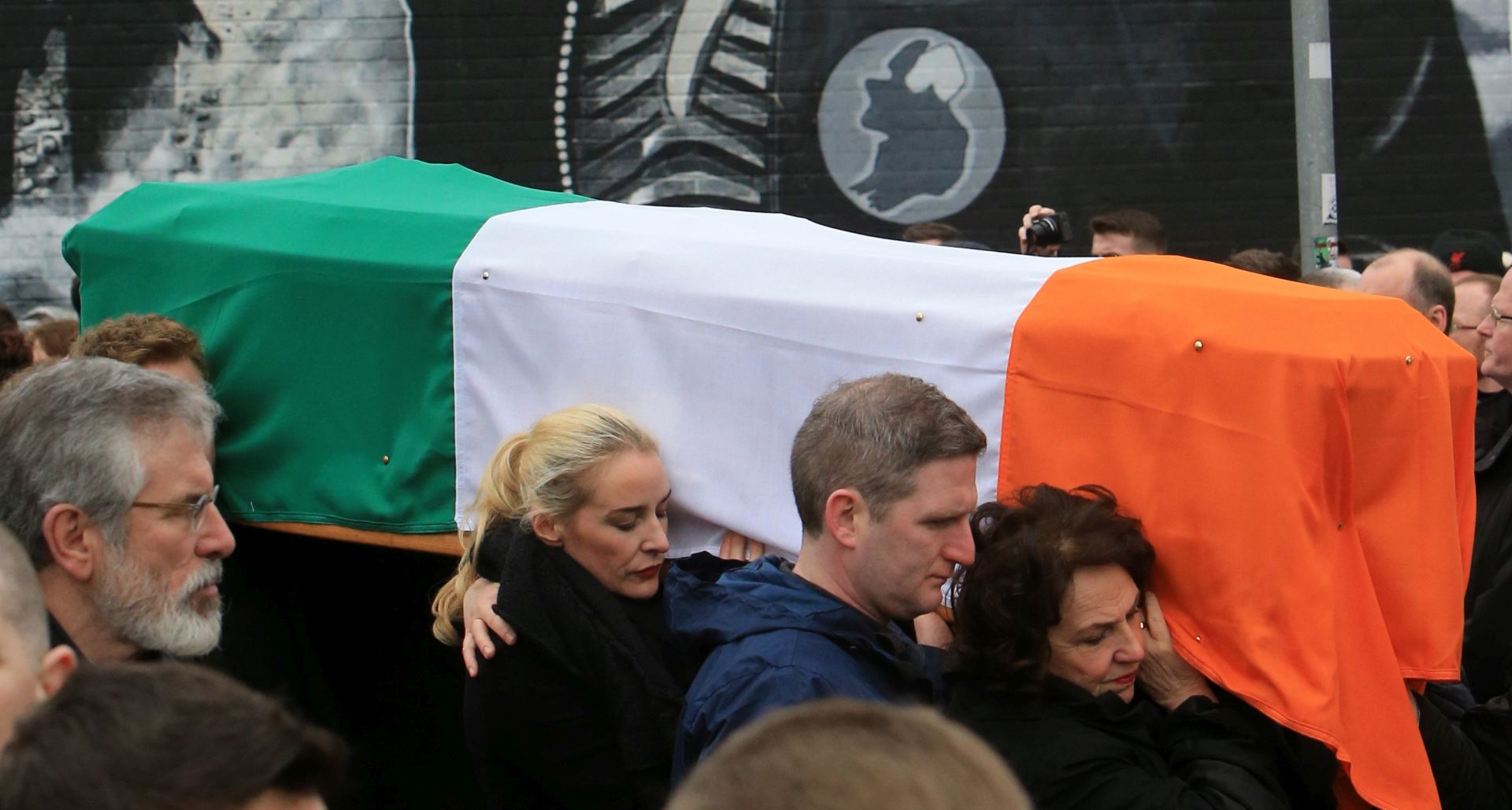 epa05865900 Bernadette Canning (R) helps carrying the coffin of her husband Martin McGuinness in front of a mural in Londonderry, Northern Ireland, 23 March 2017, during the funeral  of the late Sinn Fein leader. Sinn Fein's Martin McGuinness, Northern Ireland's former deputy first minister, died aged 66 on 21 March 2017. On far left is Sinn Fein President Gerry Adams. It is understood he had been suffering from a rare heart condition. The former IRA leader turned peacemaker worked at the heart of the power-sharing government following the 1998 Good Friday Agreement. He became deputy first minister in 2007, standing alongside Democratic Unionist Party leaders Ian Paisley, Peter Robinson and Arlene Foster.  EPA/PAUL MCERLANE