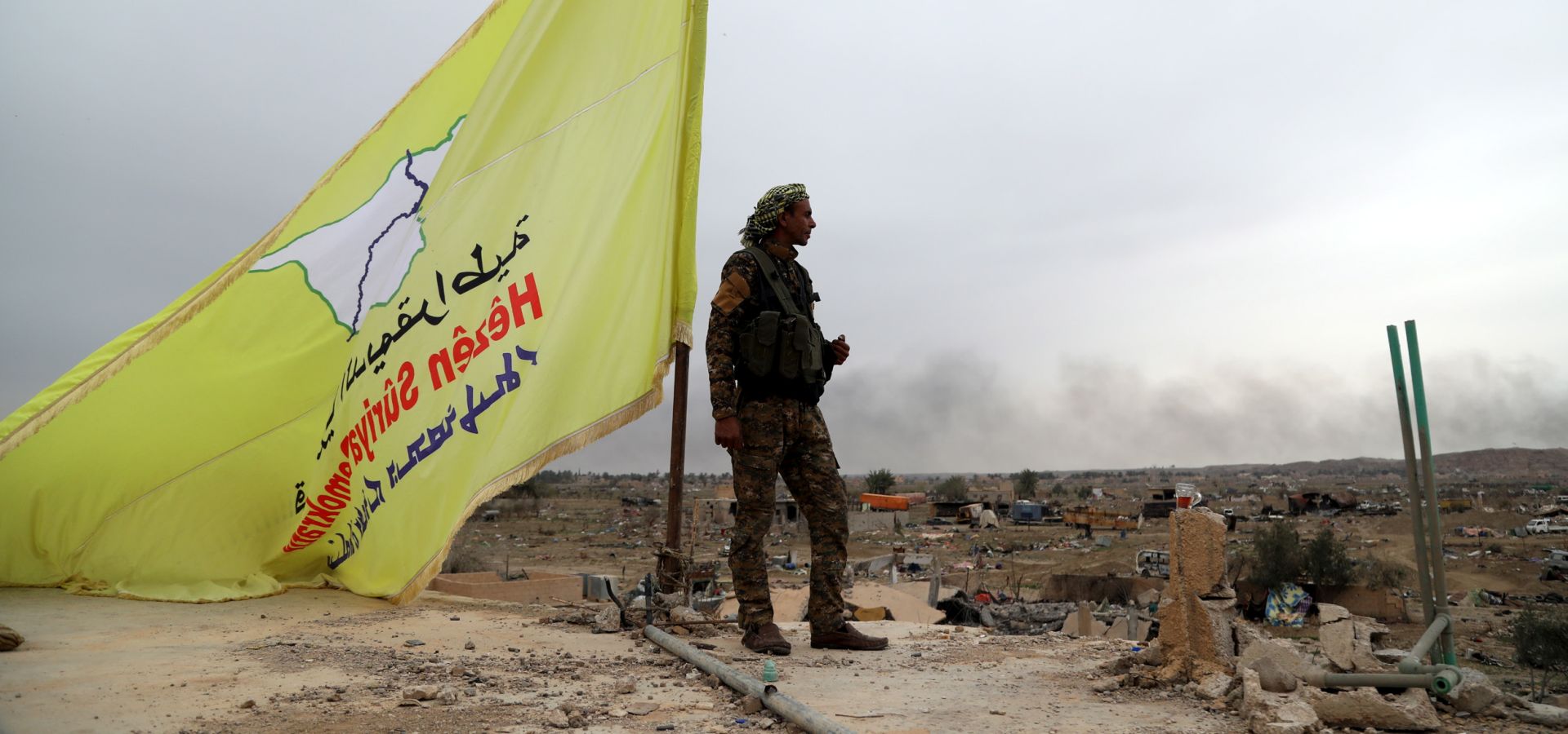 epa07461235 A Fighter of the Syrian Democratic Forces (SDF) stands guard next to the yellow flag of US-backed Syrian Democratic Forces (SDF) atop a building in the village of Baghuz, Syria, 24 March 2019. The US-backed Kurdish-Arab 'Syria democratic forces' (SDF) have announced the military victory over Islamic State (IS) group on 23 March 2019, following a four-year battle against the group that took control over a third of Syria and Iraq.  EPA/Ahmed Mardnli