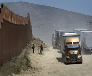 epa07626208 Two men who are fixing the road in exchange for tips watch as trucks drive along a dirt road next to the U.S.-Mexico border fence toward the commercial border inspection stationn in Tijuana, Mexico, on 04 June 2019, following US President Donald J. Trump's decision to impose tariffs on Mexico. The truck will queue in line with several hundred other trucks waiting to cross the border into San Diego, California, USA. US President Donald J. Trump stated he is planning to implement his decision to impose five percent tariffs on goods imported into the US from Mexico, with a five percent increase each month until October, if Mexican officials do not stop undocumented migrants from crossing into the US from the two nation's shared border.  EPA/David Maung