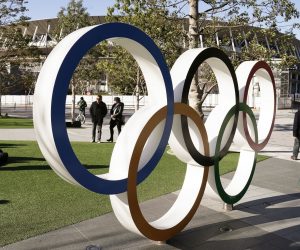 epa08251591 Visitors walk past the Olympic Rings monument in front of the Japan Olympic Committee headquarters near the new National Stadium (back), venue of the Opening and Closing Ceremony of the Tokyo 2020 Summer Olympic Games, in Tokyo, Japan, 27 February 2020. Preparations for the Tokyo 2020 Olympic Games, scheduled to start on 24 July 2020, continue as planned despite the spreading Covid-19 coronavirus outbreak, organizers confirmed.  EPA/KIMIMASA MAYAMA