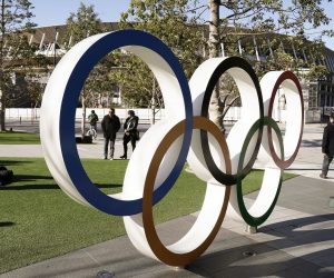 epa08251591 Visitors walk past the Olympic Rings monument in front of the Japan Olympic Committee headquarters near the new National Stadium (back), venue of the Opening and Closing Ceremony of the Tokyo 2020 Summer Olympic Games, in Tokyo, Japan, 27 February 2020. Preparations for the Tokyo 2020 Olympic Games, scheduled to start on 24 July 2020, continue as planned despite the spreading Covid-19 coronavirus outbreak, organizers confirmed.  EPA/KIMIMASA MAYAMA