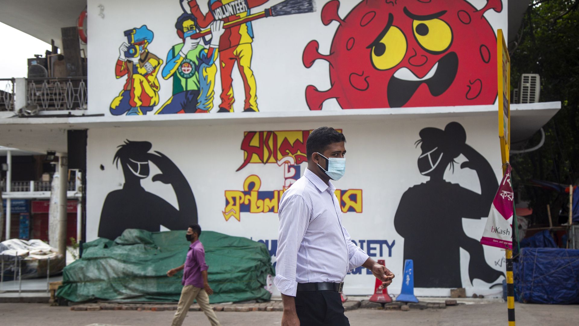 epa08513536 Bangladeshi people wearing face masks walk in front of graffiti in Dhaka, Bangladesh, 28 June 2020. Countries around the world are taking increased measures to stem the spread of the SARS-CoV-2 coronavirus which causes the COVID-19 disease.  EPA/MONIRUL ALAM