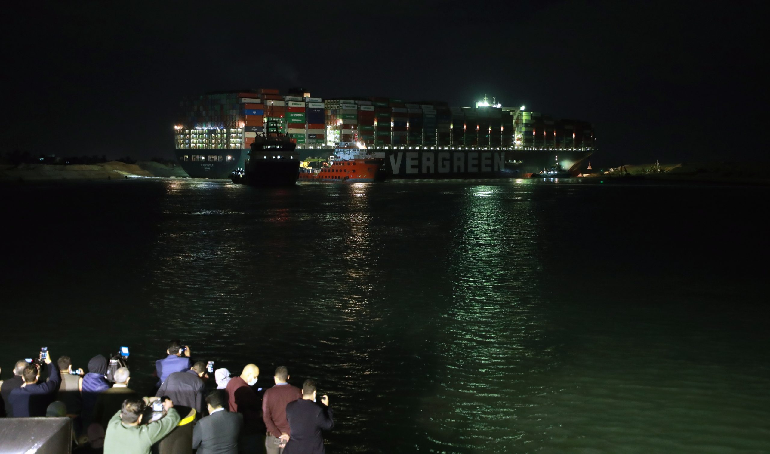 epaselect epa09101957 The Ever Given container ship which ran aground in the Suez Canal, Egypt, 27 March 2021. The Ever Given, a large container ship ran aground in the Suez Canal on 23 March, blocking passage of other ships and causing a traffic jam for cargo vessels. The head of the Suez Canal Authority announced that 'the navigation through the Suez Canal is temporarily suspended' until the floatation of the Ever Given is completed. Its floatation is being carried out by eight large tugboats that are towing and pushing the grounding vessel  EPA/KHALED ELFIQI
