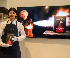 epa09993851 An art handler presents a fireproof edition of Margaret Atwood's 'The Handmaid's Tale' as a video of Atwood using a flamethrower plays on a screen behind them at the Sotheby's auction house in New York, New York, USA, 03 June 2022. Atwood and Penguin Random House partnered to make the special edition of Atwood's often banned book, up for auction online until June 7th.  EPA/SARAH YENESEL