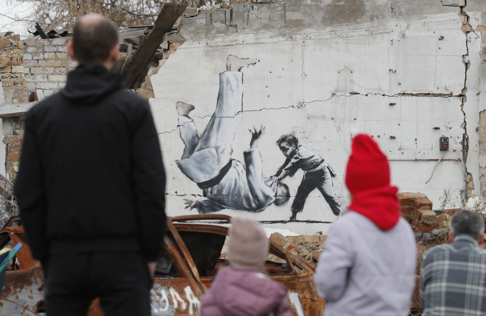 epa10303684 People watch a mural depicting child overthrowing a man in judo clothes, in a style resembling the works of British street artist Banksy, at a wall of a building that was destroyed during shelling in Borodyanka, Ukraine, 13 November 2022. A photo of a mural in Borodyanka depicting a gymnast was shared by Banksy on his social media channel on 11 November, while other works attributed to him in different locations in Ukraine and resembling Banksy's style have not been shared on the artist's channel yet. Russian troops entered Ukraine on 24 February 2022, starting a conflict that has provoked destruction and a humanitarian crisis.  EPA/SERGEY DOLZHENKO