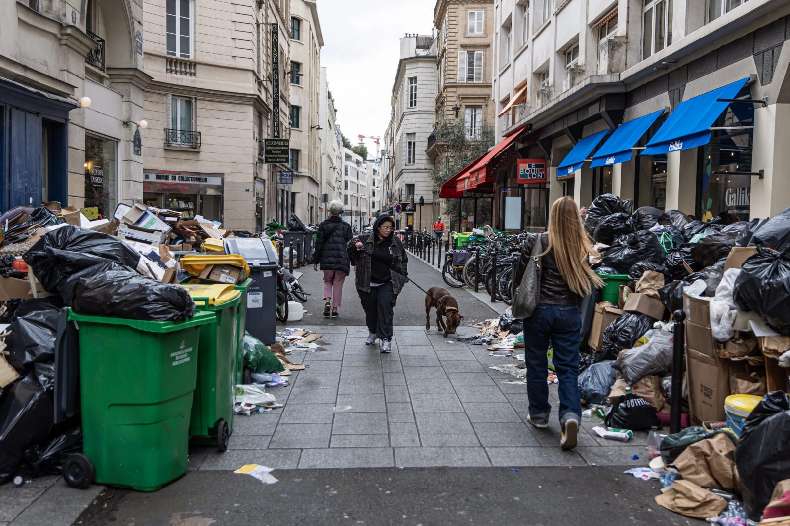 epa10535245 People pass next to garbage cans overflowing with trash in Paris, France, 21 March 2023. Parisian streets are full of more than 9,000 tons of waste yet to be collected after two weeks of garbage collectors' strike, who have joined the massive strikes in France against the government's pension reform plans after French prime minister on 16 March had announced the use of article 49 paragraph 3 (49.3) of the Constitution of France to have the text on the controversial law to be definitively adopted without a vote.  EPA/CHRISTOPHE PETIT TESSON