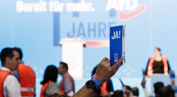 epa10773477 A delegate holds a voting card reading 'yes' as members of the counting committee walk in the background (orange vests) during the Alternative for Germany (AfD) party convention in Magdeburg, Germany, 28 July 2023. The Alternative for Germany (AfD) 14th federal party congress takes place in Magdeburg on 28 July, followed by a five-day European Elections assembly.  EPA/CLEMENS BILAN