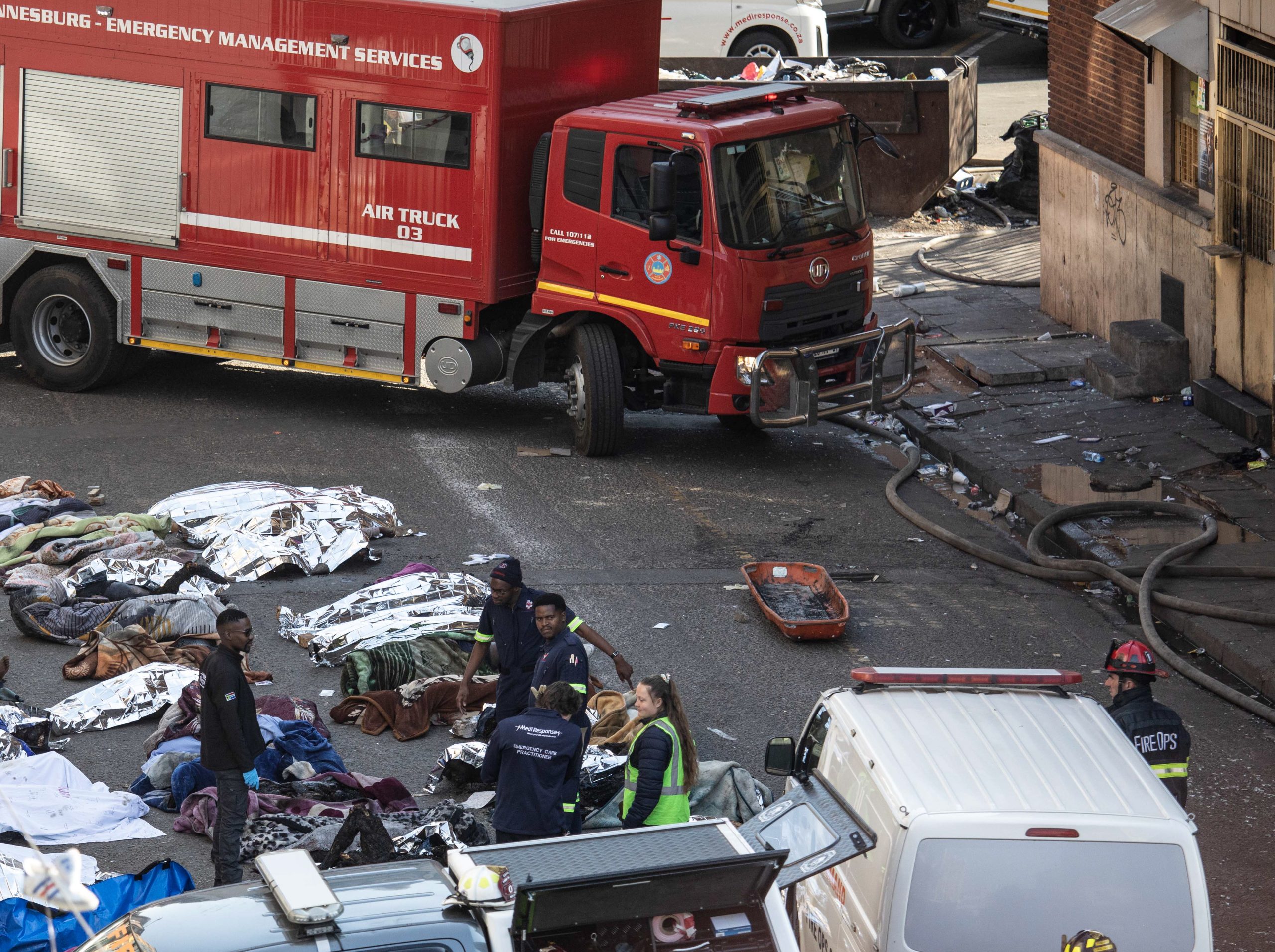 epa10830422 Fire crews and emergency staff stand next to the covered bodies of the victims as they gather at the site of a fire that broke out at the five-storey building in the city centre, in Johannesburg, South Africa, 31 August 2023. More than 60 people have died following a fire that ravaged a building in central Johannesburg according to officials.  EPA/KIM LUDBROOK