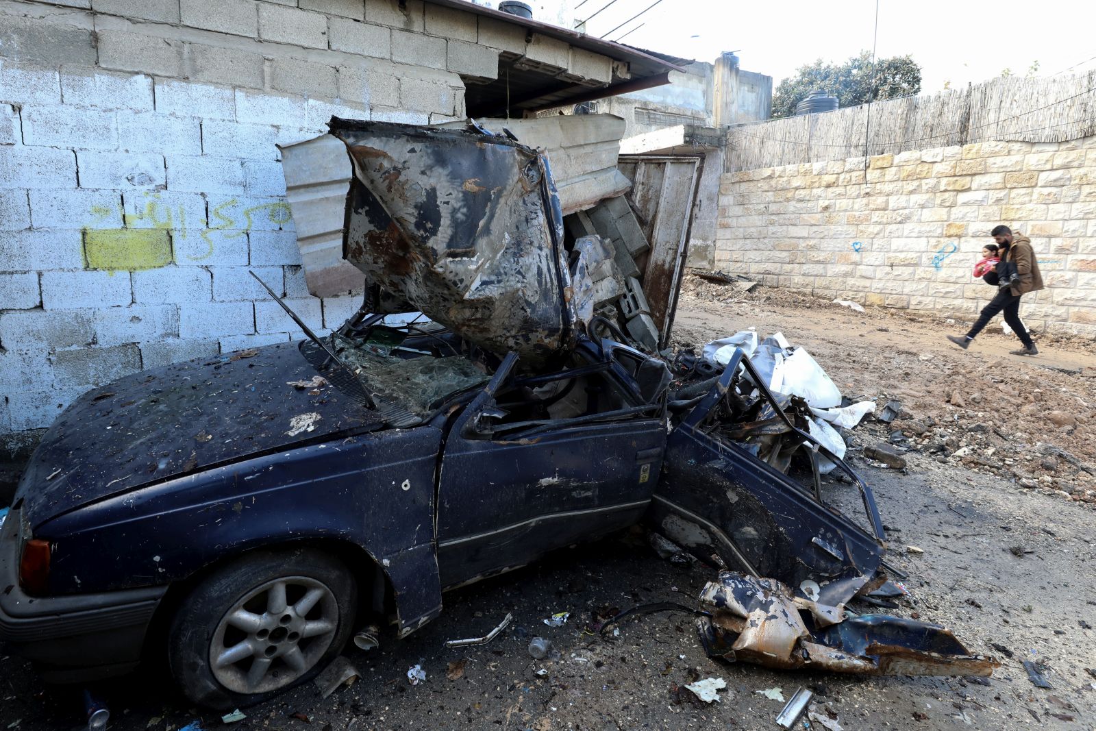 epaselect epa11076308 A Palestinian man carrying a child walks past a damaged car at the Jenin refugee camp following an overnight Israeli raid in the West Bank city of Jenin, 14 January 2024. Israeli forces stormed the city of Jenin and its camp on 13 January evening, provoking confrontations with local residents, according to the Palestinian news agency Wafa.  EPA/ALAA BADARNEH