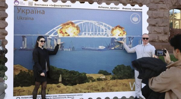 People pose in Kyiv on Oct. 8, 2022, for a photo in front of an oversized stamp featuring an artist's impression of an explosion earlier that day which severely damaged the Kerch Bridge linking mainland Russia to the Russian-annexed Crimean Peninsula. (Kyodo) ==Kyodo  Photo via Newscom