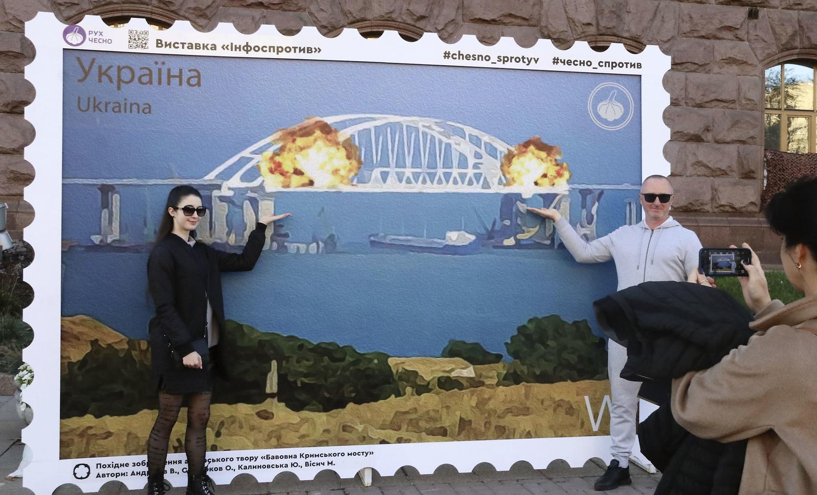 People pose in Kyiv on Oct. 8, 2022, for a photo in front of an oversized stamp featuring an artist's impression of an explosion earlier that day which severely damaged the Kerch Bridge linking mainland Russia to the Russian-annexed Crimean Peninsula. (Kyodo) ==Kyodo  Photo via Newscom
