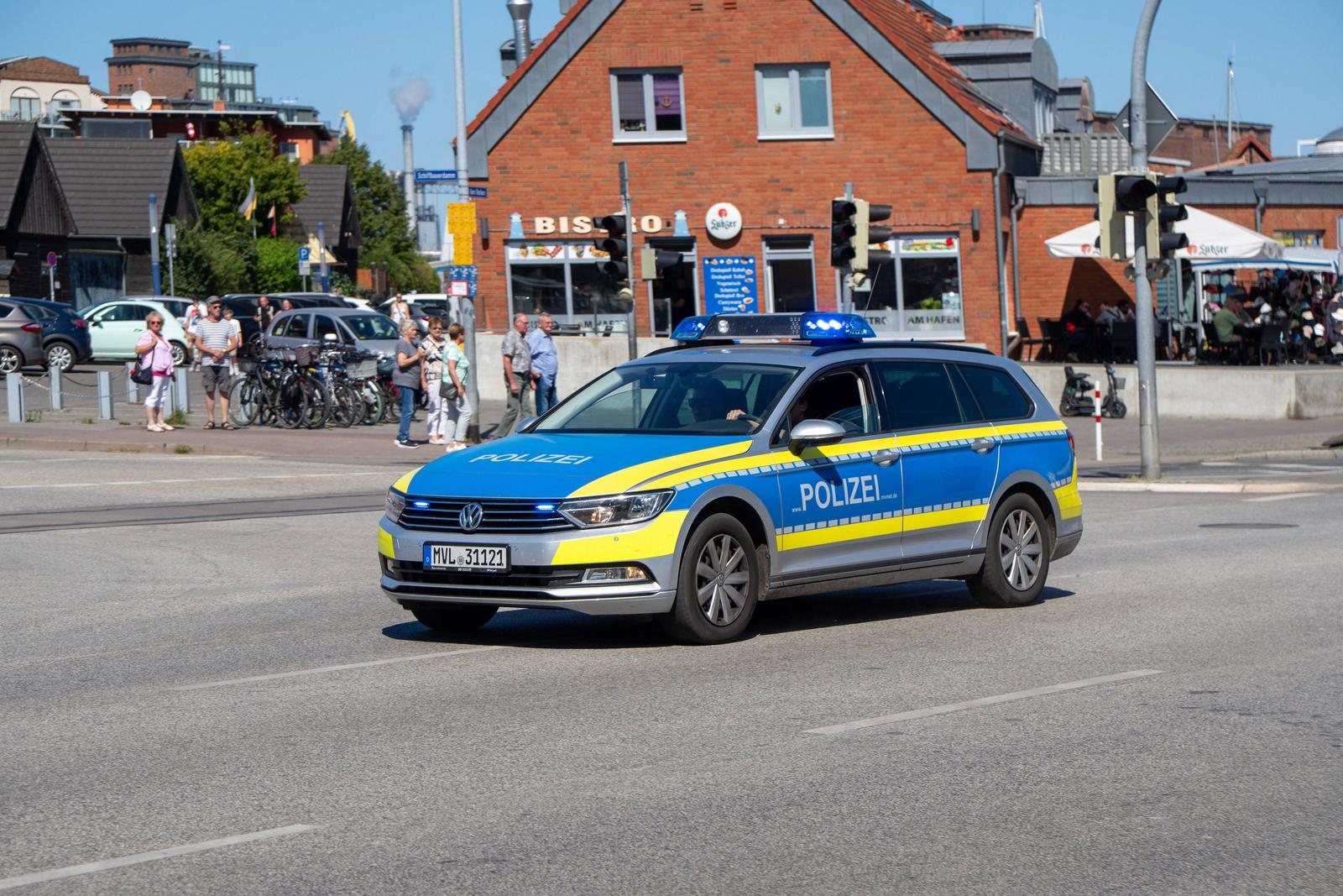 Wismar, Deutschland 19.Aug.2025: Im Bild: Polizeifahrzeug Volkswagen Passat Variant mit blauer und gelber Lackierung und eingeschaltetem Blaulicht im Einsatz in der Innenstadt von Wismar, Polizei Mecklenburg-Vorpommern, Sicherheitskräfte im Streifendienst, sichtbare Polizeipräsenz im Stadtverkehr, Verkehrssicherheit, Kontrolle, Einsatz gegen Kriminalität, Streifenwagen vor Bistro am Hafen und Passanten im Hintergrund. Mecklenburg Vorpommern *** Wismar, Germany 19 Aug 2025 In the picture police car Volkswagen Passat Variant with blue and yellow paintwork and switched on blue light in action in the city center of Wismar, police Mecklenburg Vorpommern, security forces on patrol duty, visible police presence in city traffic, traffic safet Copyright: xFotostandx/xGelhotx Photo: IMAGO/Fotostand / Gelhot/IMAGOSTOCK&PEOPLE